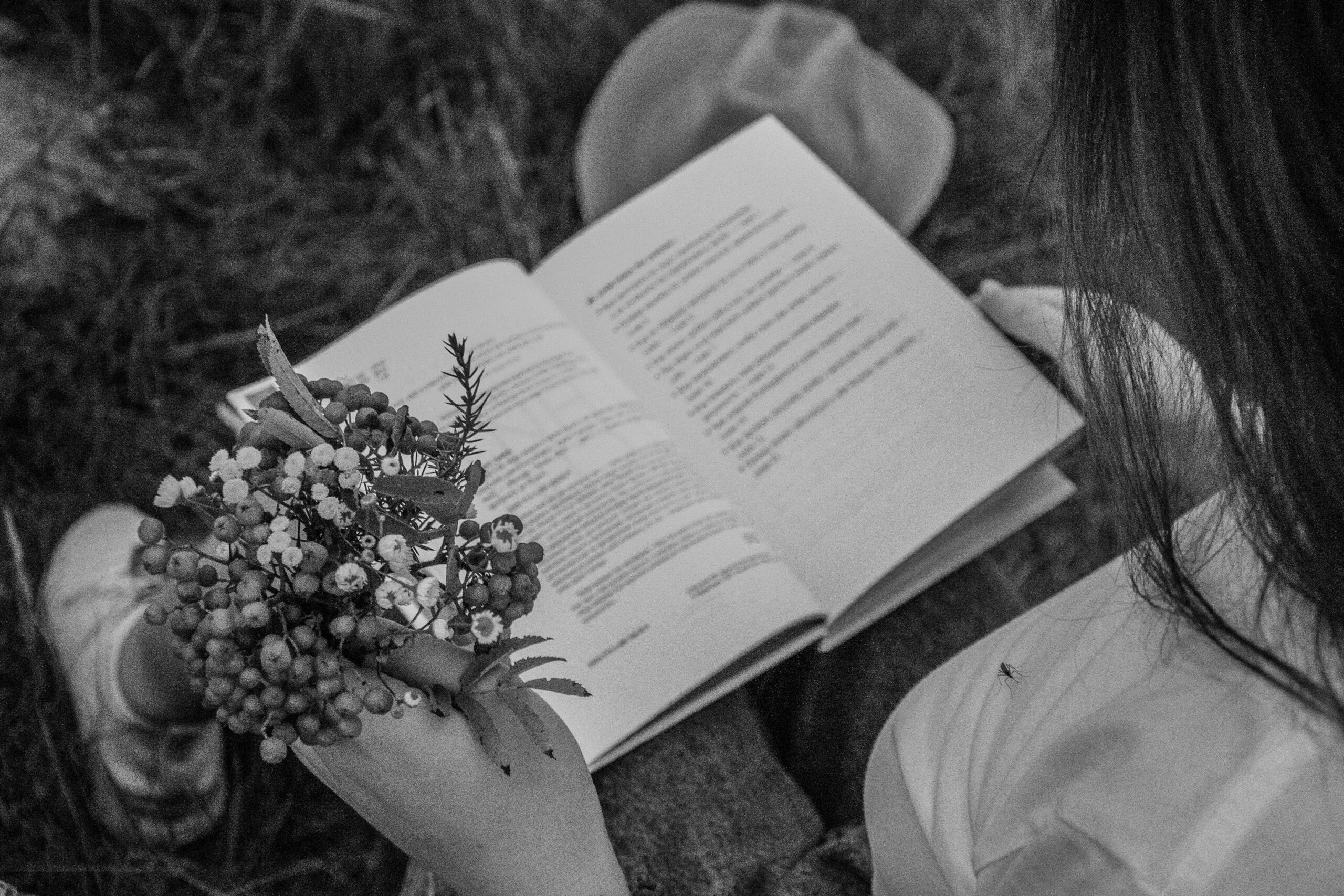 A person reads a book while holding flowers, captured in black and white outdoors.