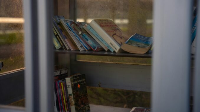A collection of books displayed on a sunlit bookshelf inside a glass enclosure.