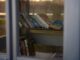 A collection of books displayed on a sunlit bookshelf inside a glass enclosure.