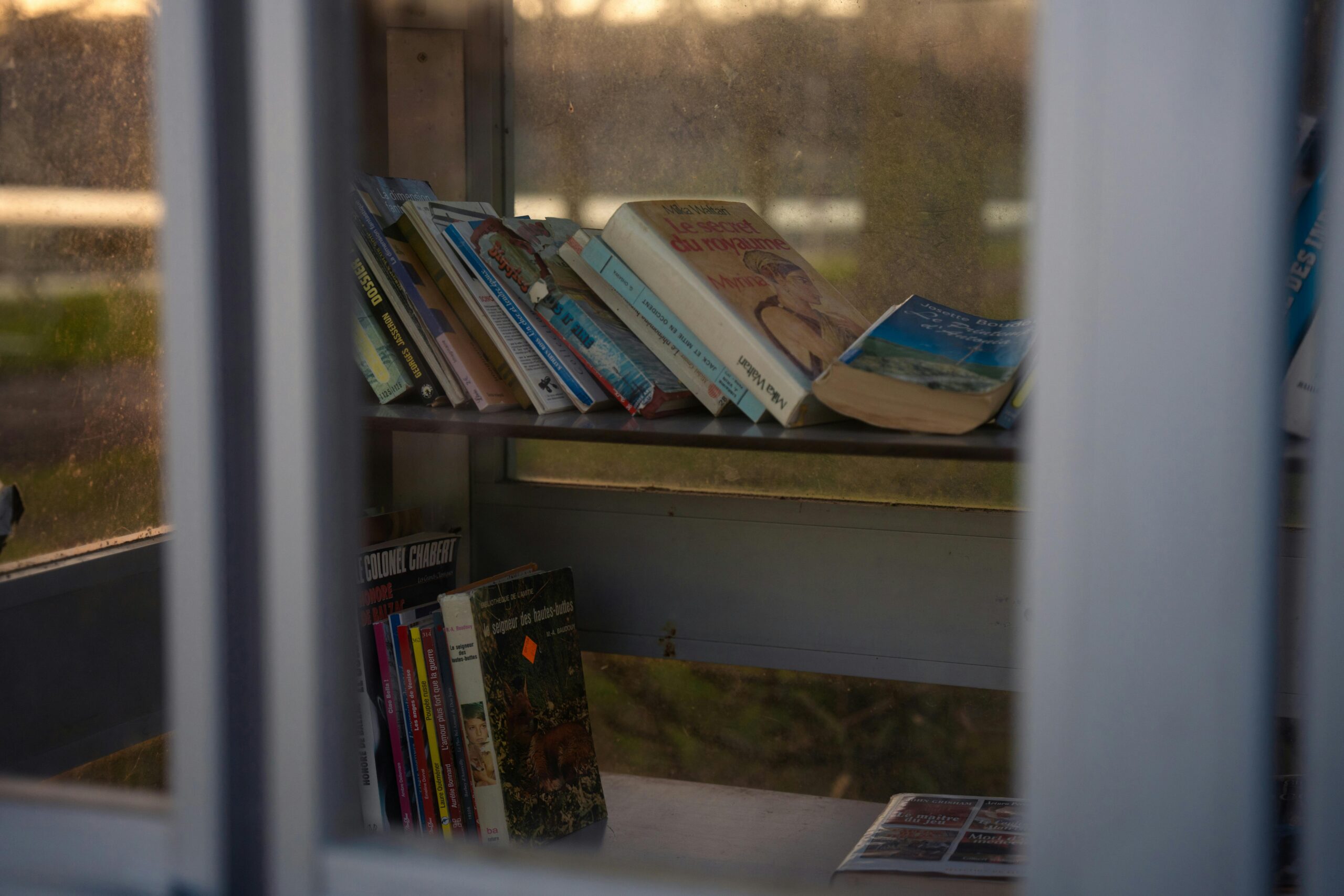 A collection of books displayed on a sunlit bookshelf inside a glass enclosure.