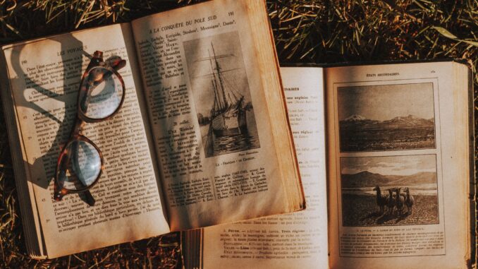 Top view paged of worn out textbooks with pictures and letters placed on grassy ground with eyeglasses in rural area