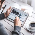 A man uses a tablet to browse news while enjoying a cup of coffee, stylus in hand.